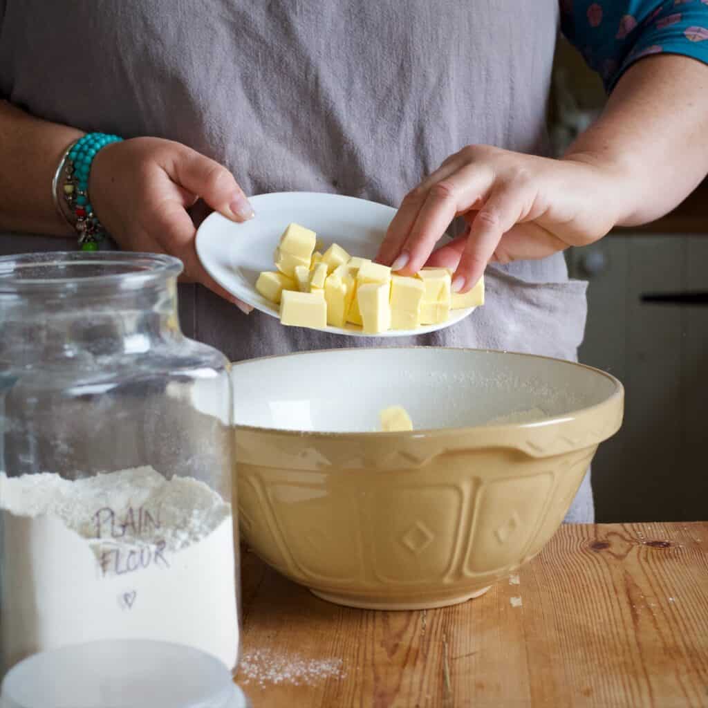 Womans hands scraping cubes of butter from a small white plate into a large brown mixing bowl