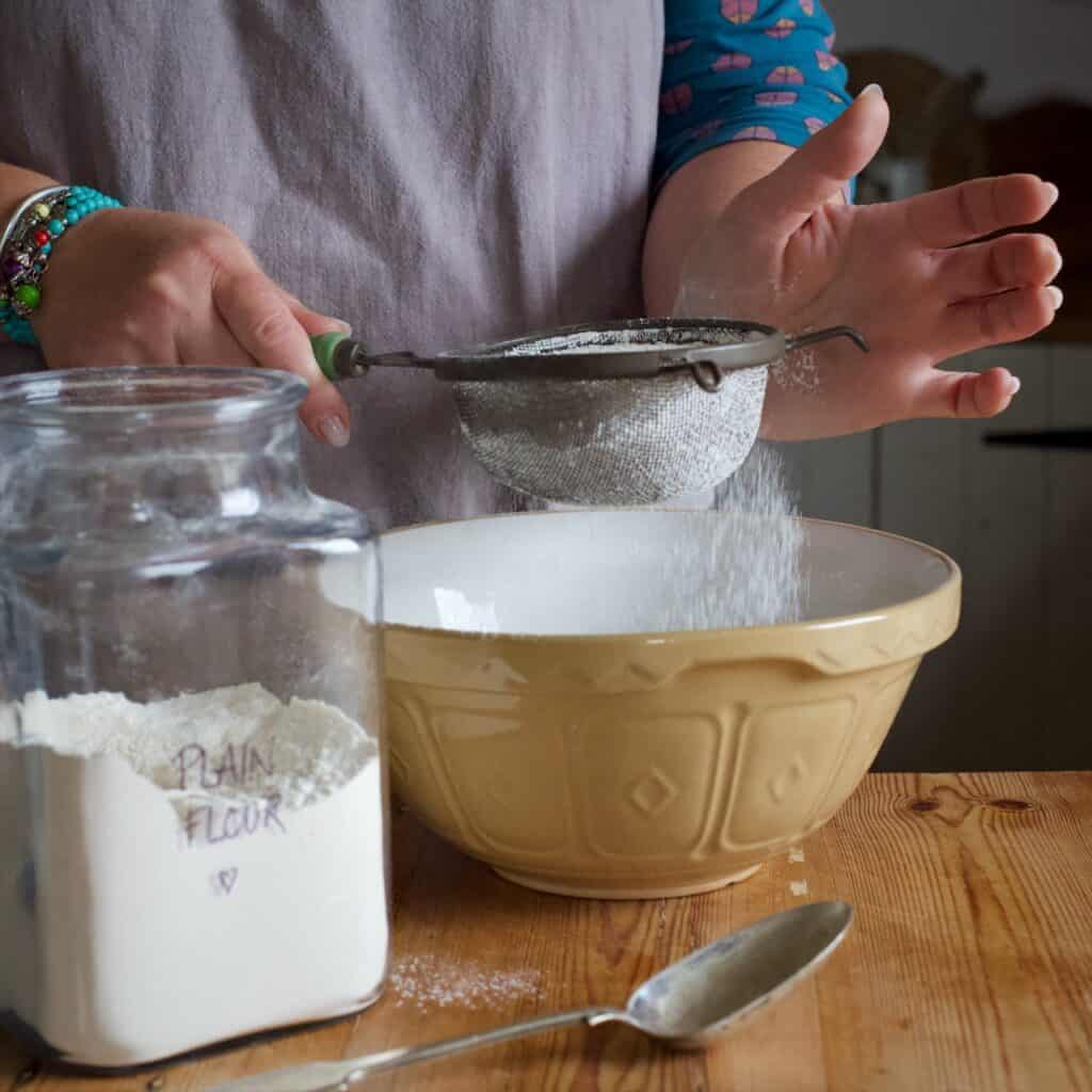 Woman in grey sieving flour from an old fashioned sieve into a brown mixing bowl