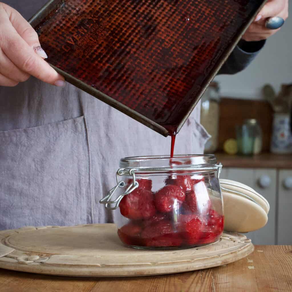 Woman in grey pouring the juice from a baking tray of roast strawberries into a glass jar filled with them