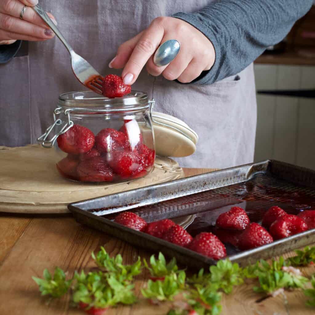 Womans hands placing cooled, roasted strawberries into a glass jar with a silver fork