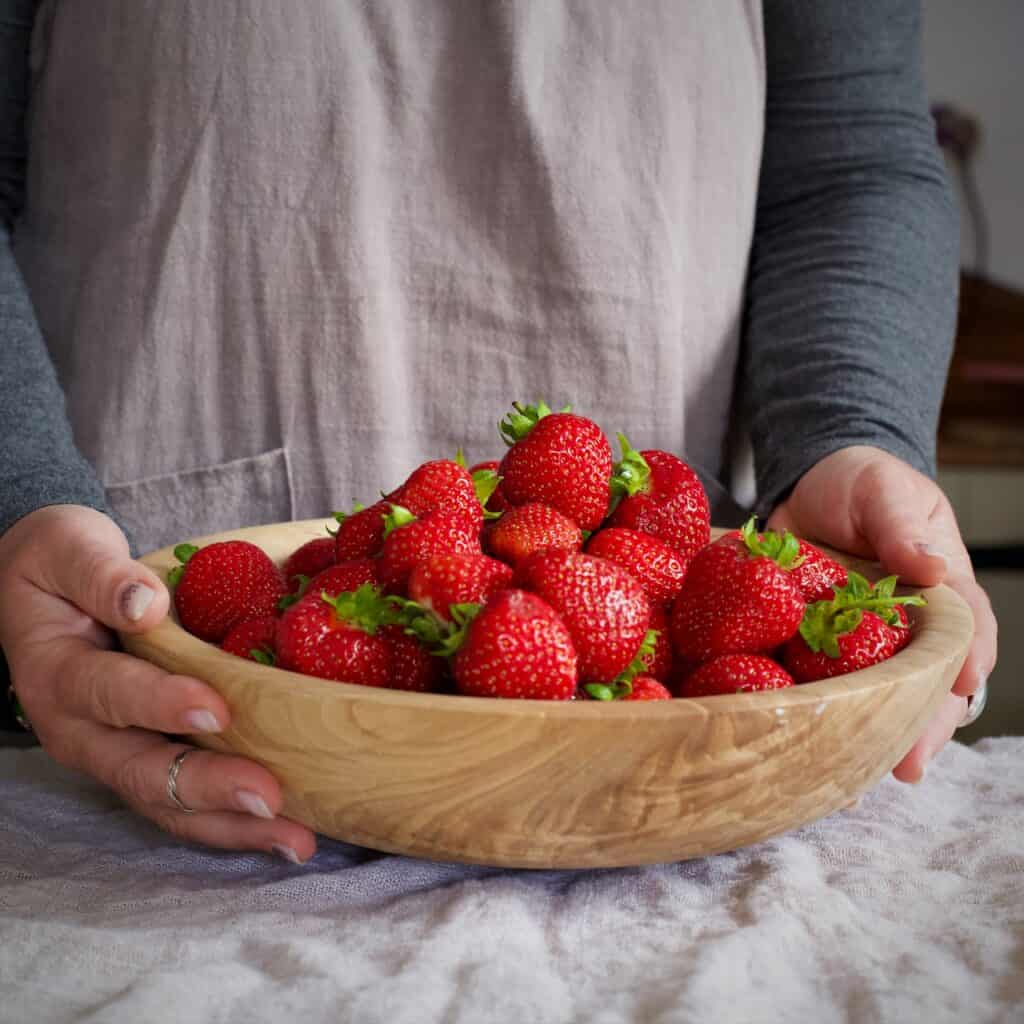 Woman in grey holding a wooden bowl filled with fresh summer strawberries