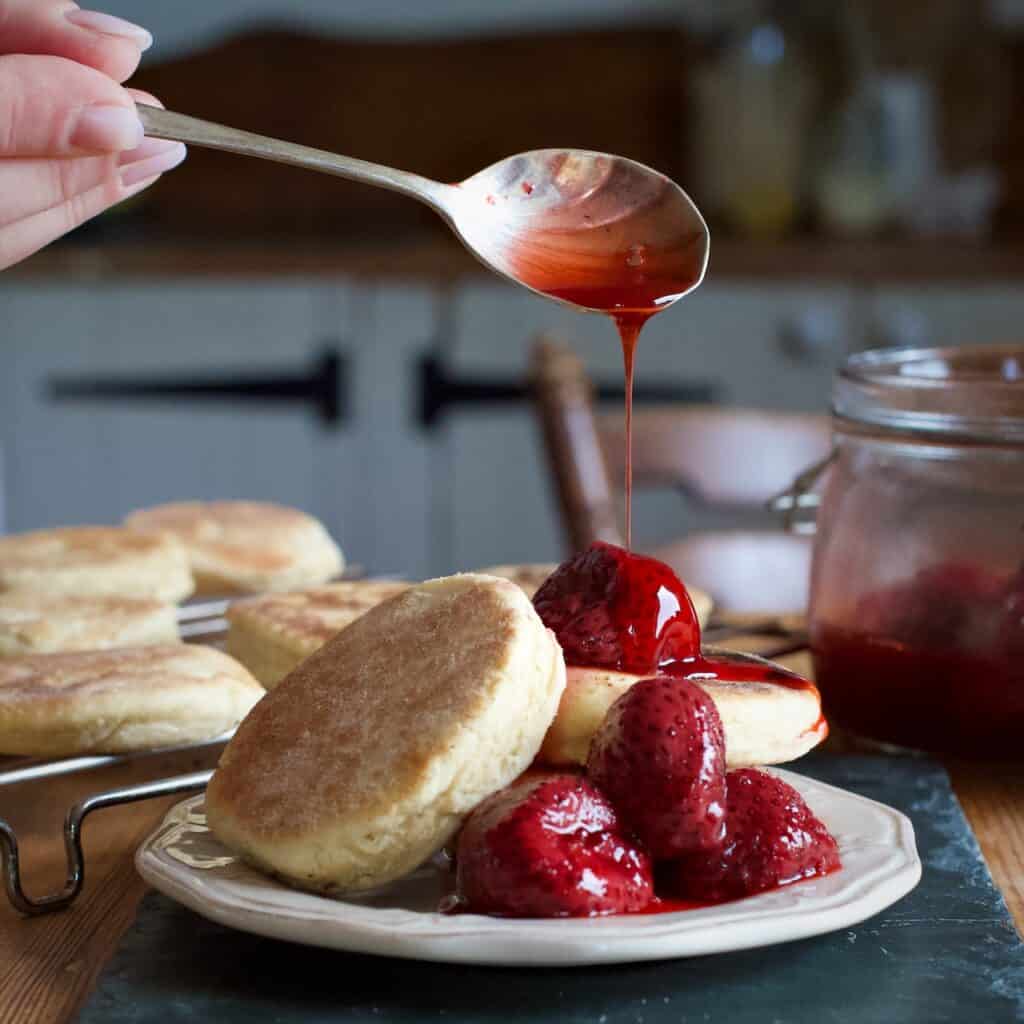 Spoon of roast strawberry juice being drizzled over a plate of griddle cakes and cooked strawberries