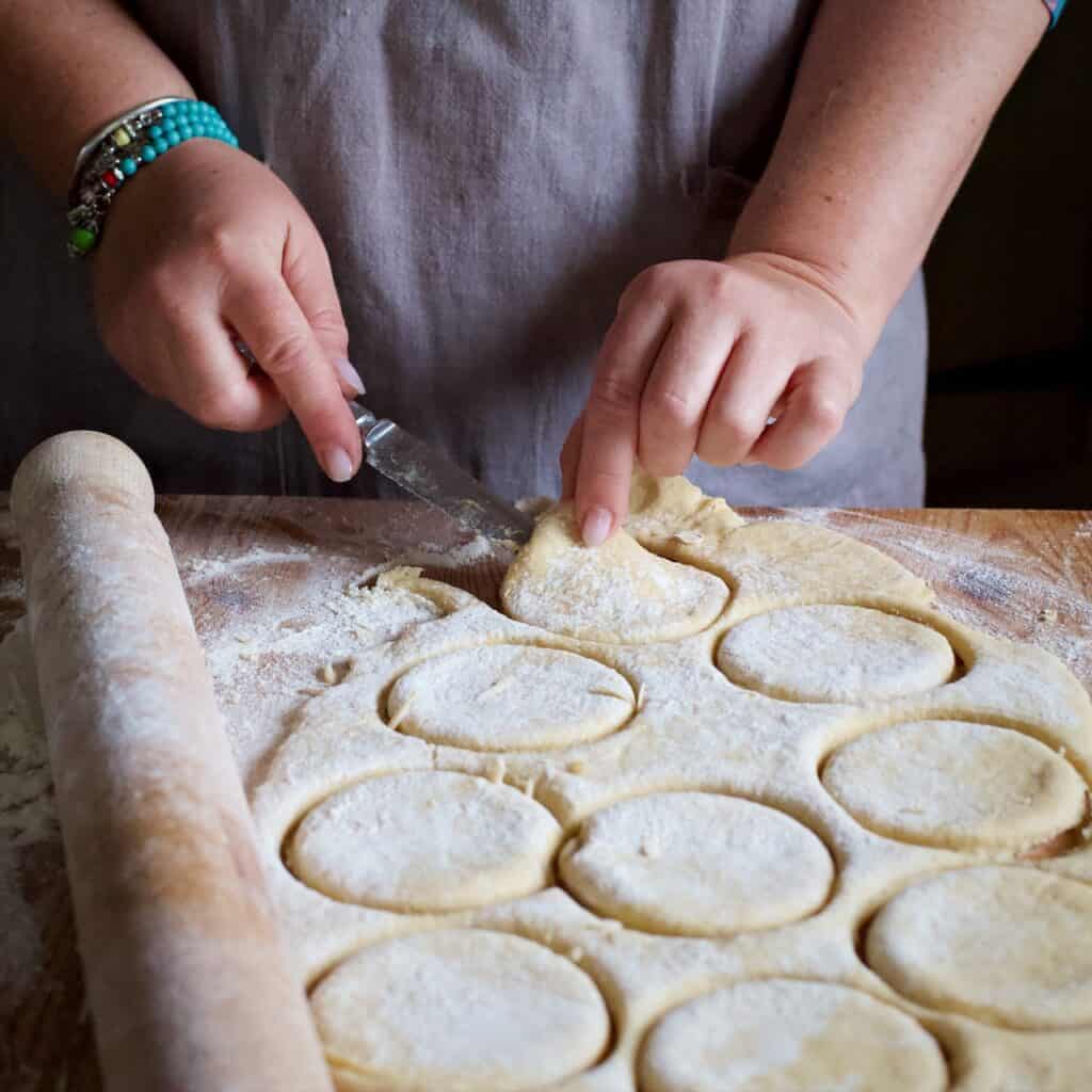 Woman in grey licking up a round of soft griddle cake dough using a silver butter knife