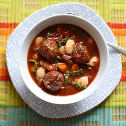 Slow Cooker Spiced Meatball Stew in a white bowl with a soup spoon on a blue and white checked plate with a striped cloth below