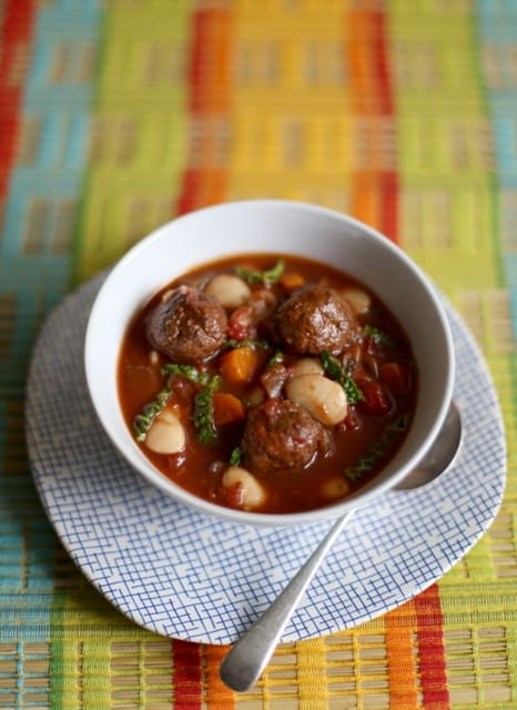 Slow Cooker Spiced Meatball Stew in a white bowl with a soup spoon on a blue and white checked plate with a striped cloth below