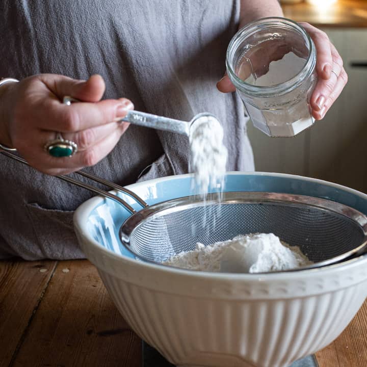 womans hands tipping baking powder into a sieve over a pale blue mixing bowl