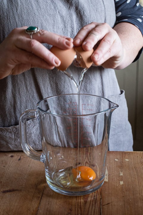 womans hands cracking an egg into a glass jug on a wooden kitchen counter