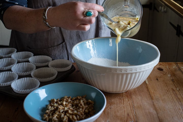 womans hands pouring wet ingredients from a glass jug into a blue mixing bowl of flour