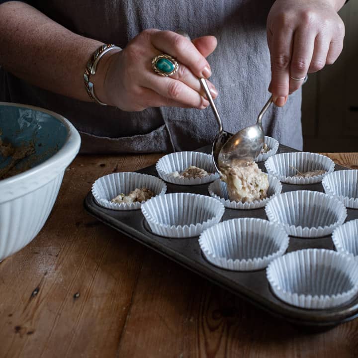 womans hands spooning pear muffin mixture into white paper muffin cases in a silver metal muffin tray