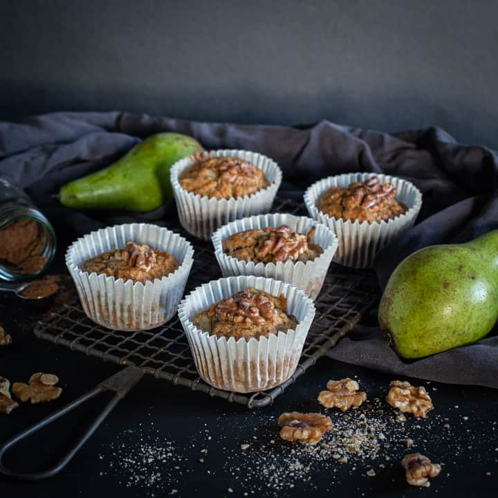 black background and 5 pear muffins on a rustic cooling rack with raw green pears and walnut halves scattered around