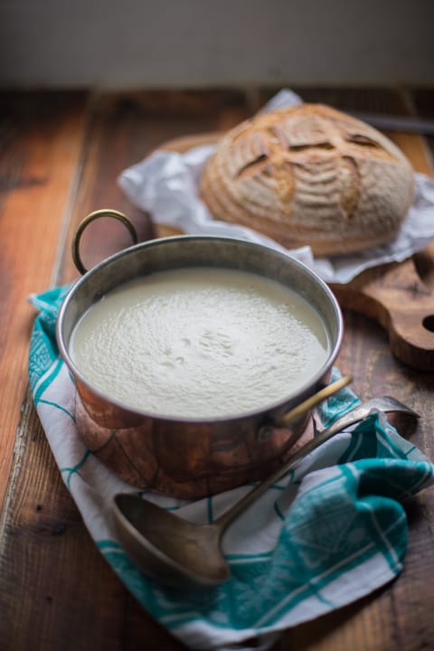 Cream of Celeriac Soup in a copper soup pan with a ladle and a round loaf on a bread board all on a wooden table