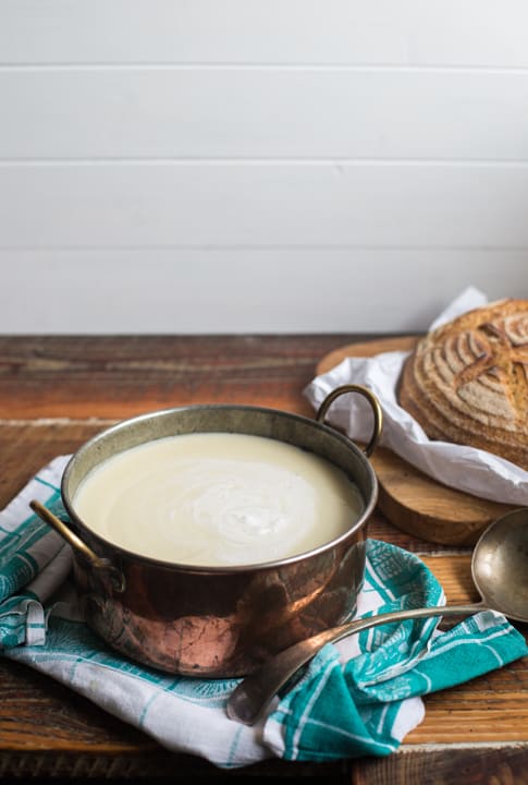 Cream of Celeriac Soup in a copper soup pan with a ladle and a round loaf on a bread board all on a wooden table