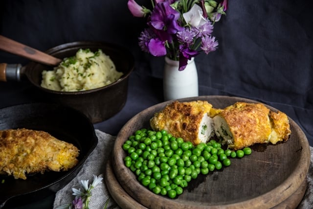 Wild Garlic Chicken Kiev on a wooden plate with green peas on a purple and lillac cloth with serving bowl of mashed potatoes and flowers in a vase