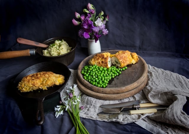 Wild Garlic Chicken Kiev on a wooden plate with green peas on a purple and lillac cloth with serving bowl of mashed potatoes and flowers in a vase