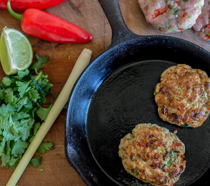 Turkey and Lemongrass Burger in a frying pan on a wooden table top surrounded by uncooked burgers ,chillis,lemongrass and lemon