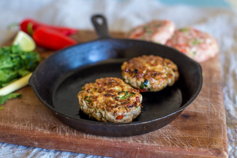 Turkey and Lemongrass Burger in a frying pan on a wooden table top surrounded by uncooked burgers ,chillis,lemongrass and lemon