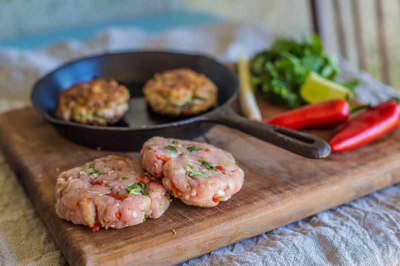 Turkey and Lemongrass Burger in a frying pan on a wooden table top surrounded by uncooked burgers ,chillis,lemongrass and lemon