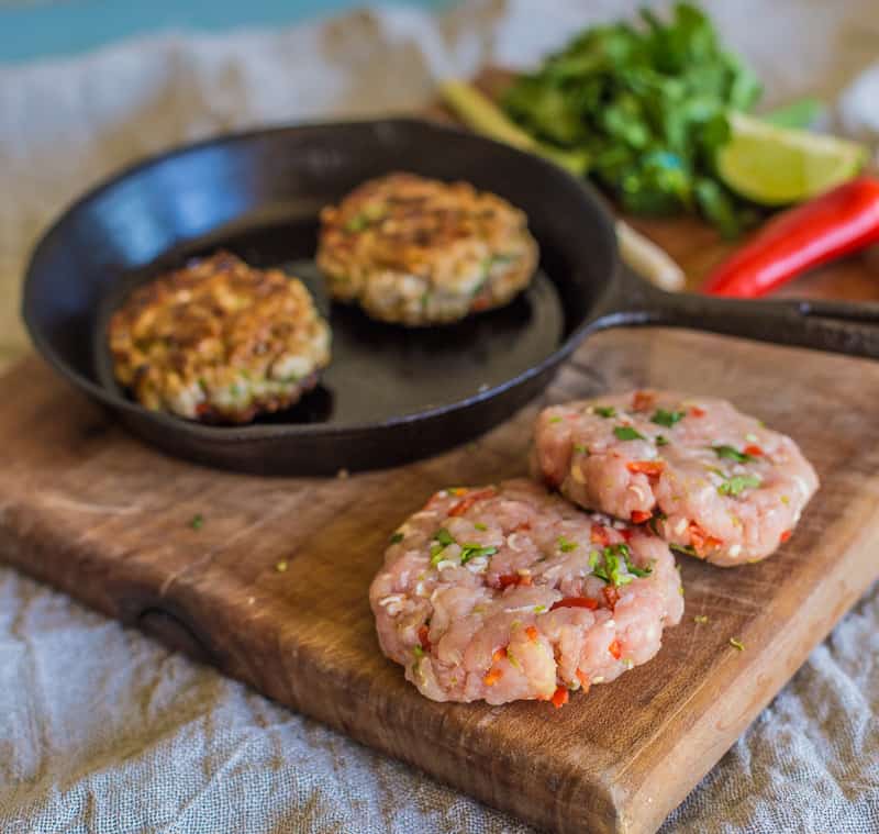 Turkey and Lemongrass Burger in a frying pan on a wooden table top surrounded by uncooked burgers ,chillis,lemongrass and lemon