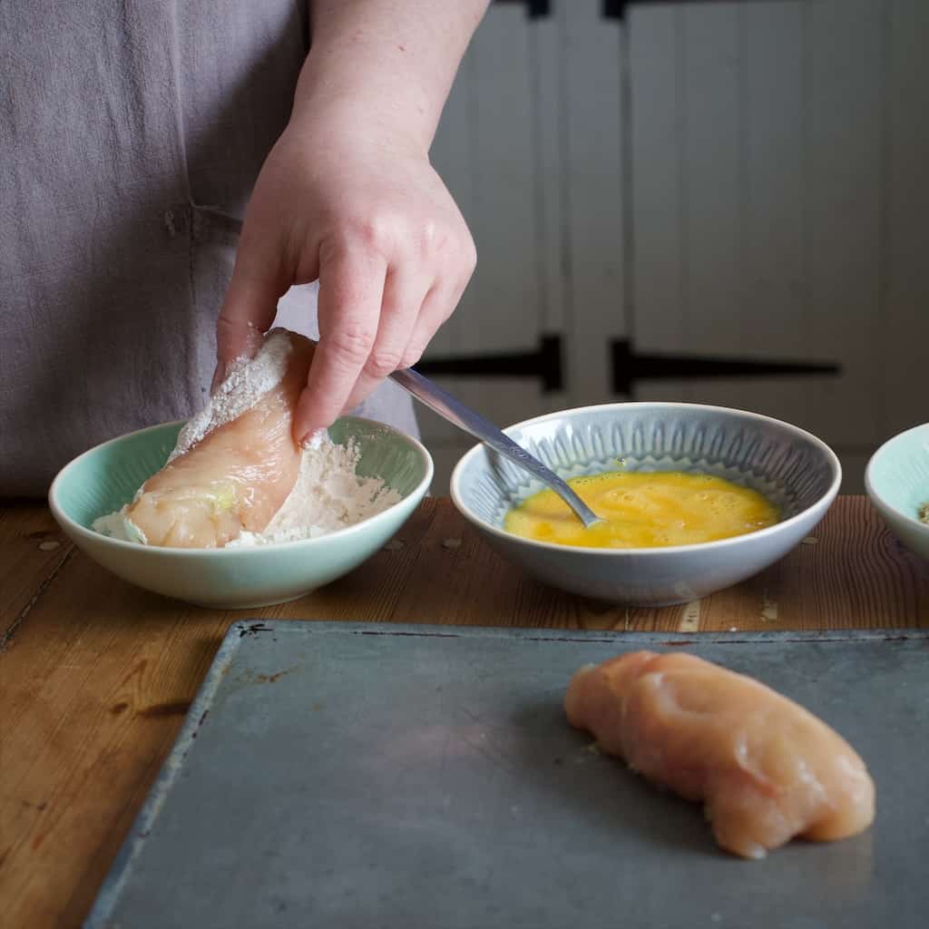 Woman in grey rolling a rolled chicken breast in a small bowl of flour before coating in breadcrumbs and baking