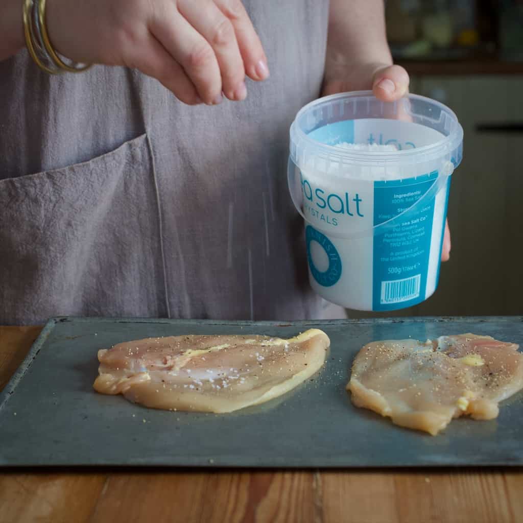 Woman sprinkling Cornish sea salt onto two chicken breast on a silver baking sheet