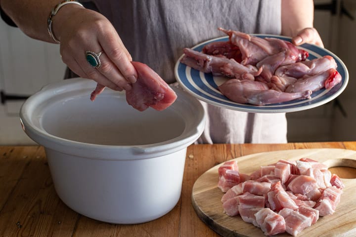 womans hands placing rabbit pieces into a white slow cooker bowl on a wooden kitchen counter