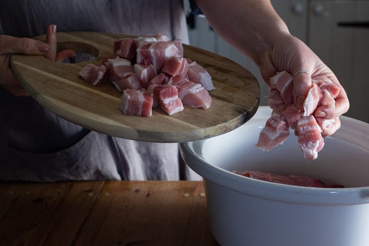 womans hands placing pork pieces into a white slow cooker bowl on a wooden kitchen counter