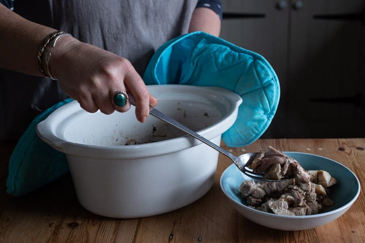 womans hands spoonign cooked rabbit from a white slow cooker into a blue bowl on a wooden kitchen counter