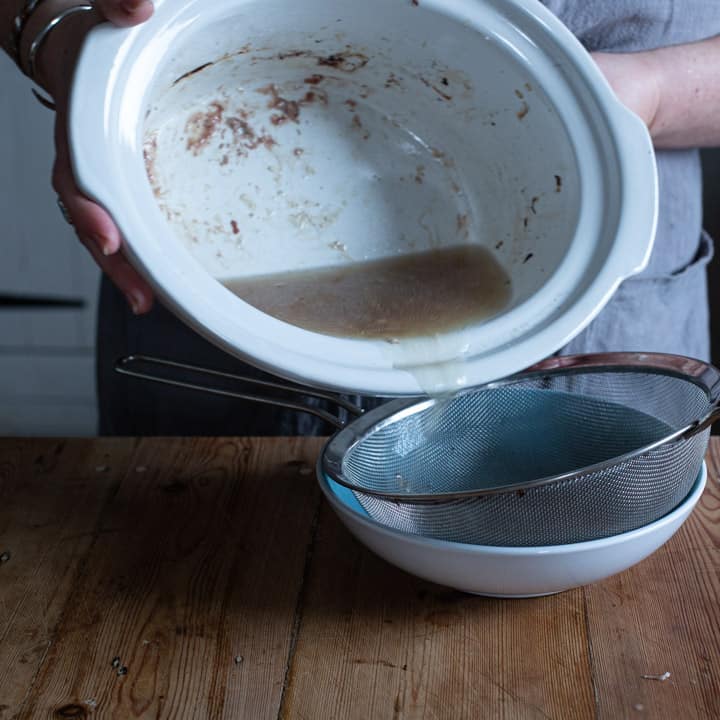 womans hands pouting rabbit stock from a white slow cooker bowl into a sieve on a wooden kitchen counter