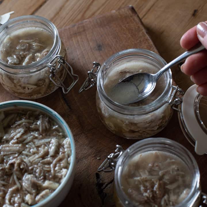 wooden background with small glass jar filled with rabbit rillettes. a small silver spoon is pushing the rabbit meat under the surface of the cooking juices to preserve it