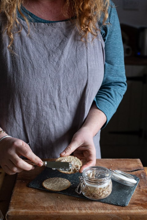 woman in grey apron spreading homemade rabbit pate on an oat cake on a wooden kitchen counter