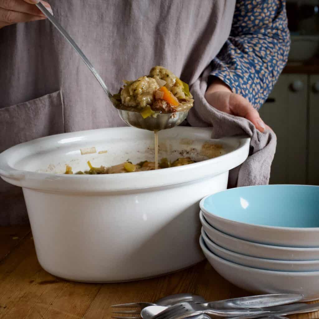 Woman ladling cooked mutton stew from a white slow cooker bowl into a blue serving bowl