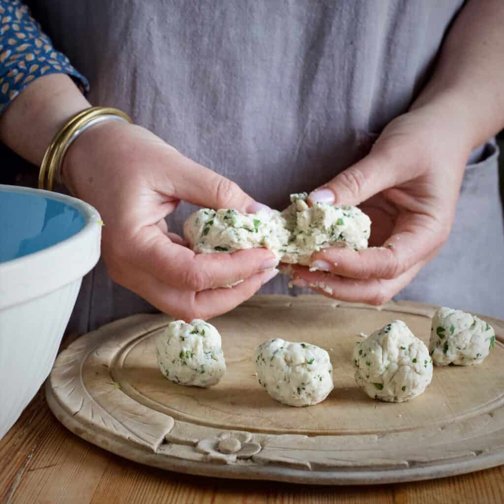 Woman splitting a piece of dumpling dough over a wooden board with 4 round dumplings on ready for cooking