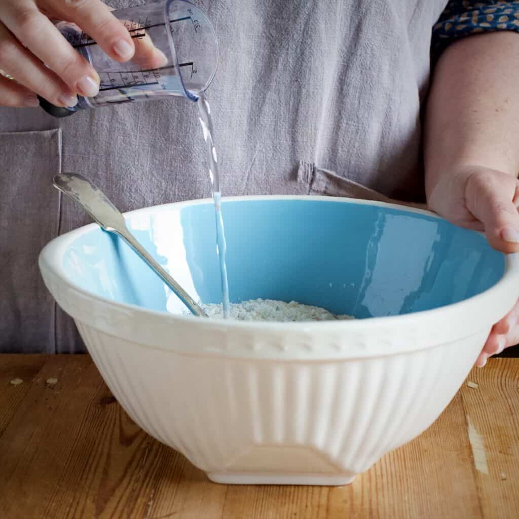 Woman pouring water from a plastic mess au ring cup into a large blue and white mixing bowl