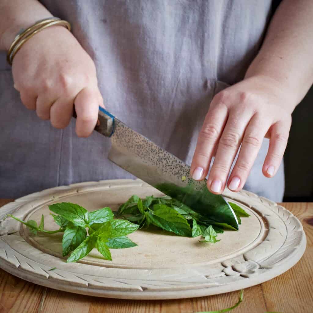 Woman in grey chopping fresh mint leaves on an old fashioned wooden chopping board