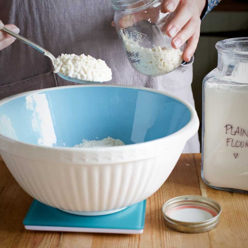 Woman spooning beef suet from a glass jar into blue and white mixing bowl