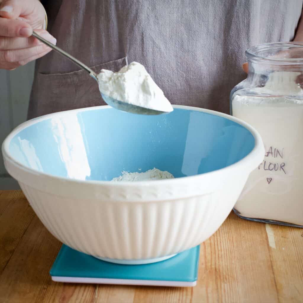 Woman spooning flour from a large glass jar into a blue and white mixing bowl