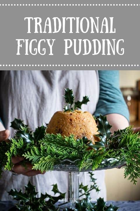 Woman’s in grey holding a a glass cake stand with green holly and leaves and a golden brown figgy pudding in the centre