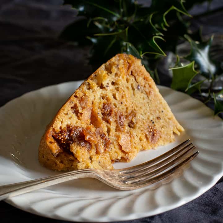A slice of golden figgy pudding on a white plate surrounded by holly leaves