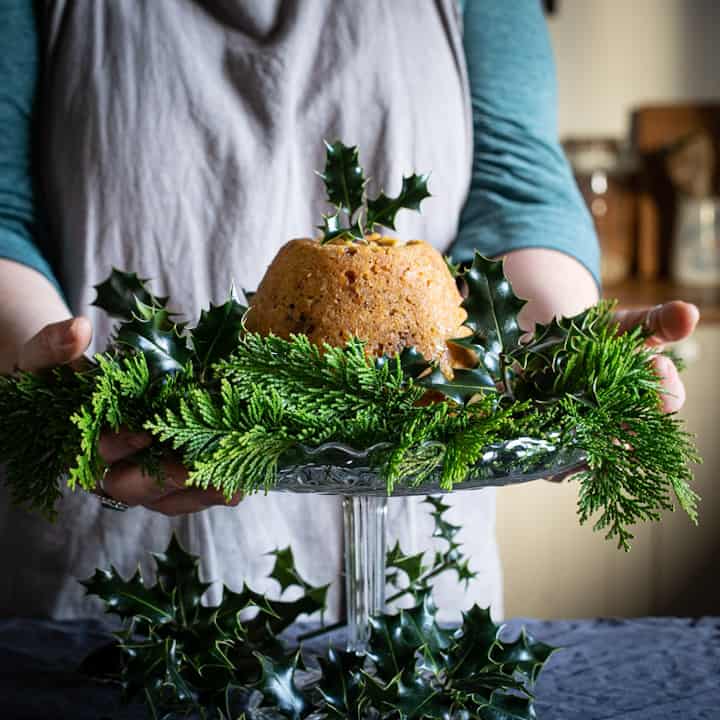 Woman’s in grey holding a a glass cake stand with green holly and leaves and a golden brown figgy pudding in the centre