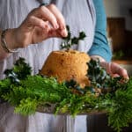 Woman’s hands placing a sprig of green holly onto a steamed pudding surrounded with holly and green foliage