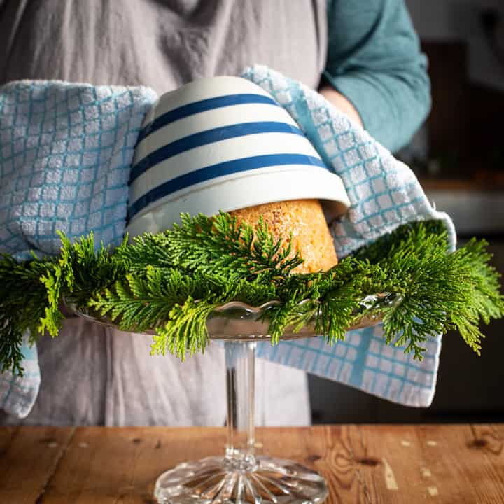 Woman’s hands turning a steamed pudding out of a blue and white pudding basin onto a glass cake stand covered in green holly and leaves