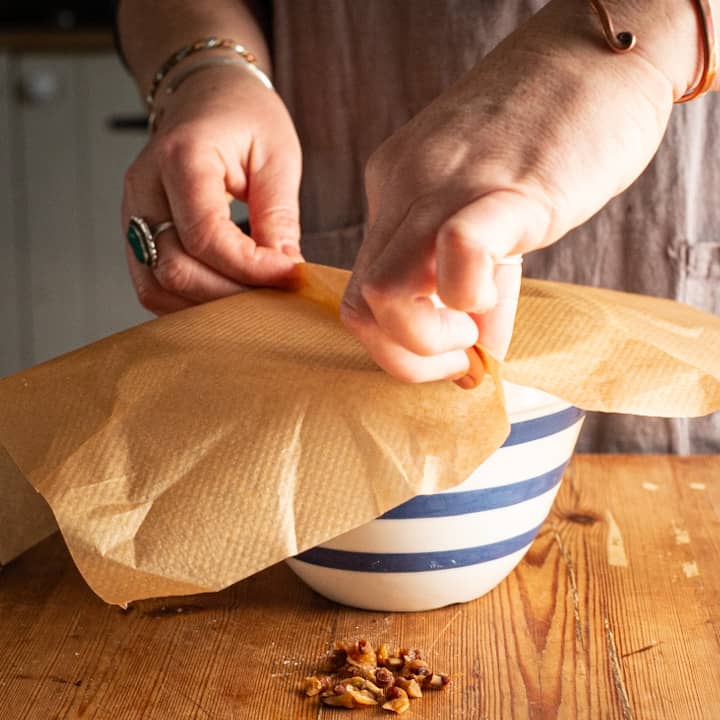 Woman’s hands pleating a piece of brown baking paper over a blue and white pudding basin on a wooden kitchen counter