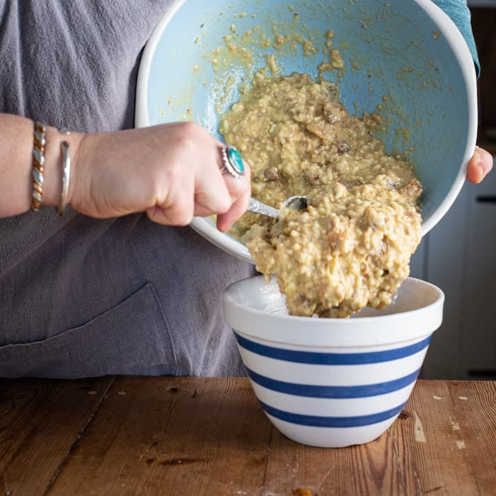 Woman scraping figgy pudding batter from a large blue mixing bowl in a stripy pudding basin on a wooden counter