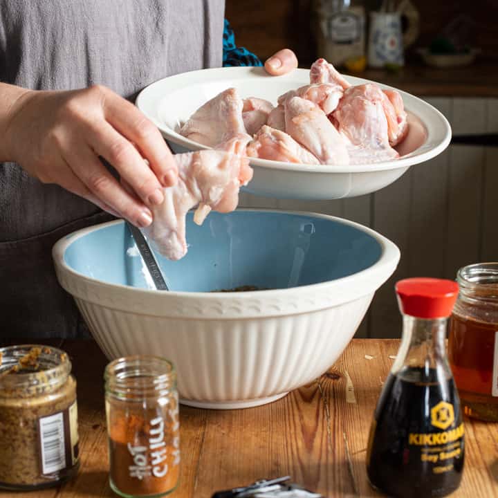 woman in grey moving raw chicken wings from a large white plate into a pale blue mixing bowl