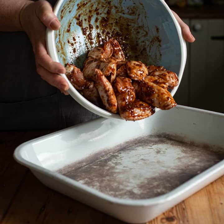 womans hands tipping a bowl of marinated chicken wings into an old white enamel baking tray