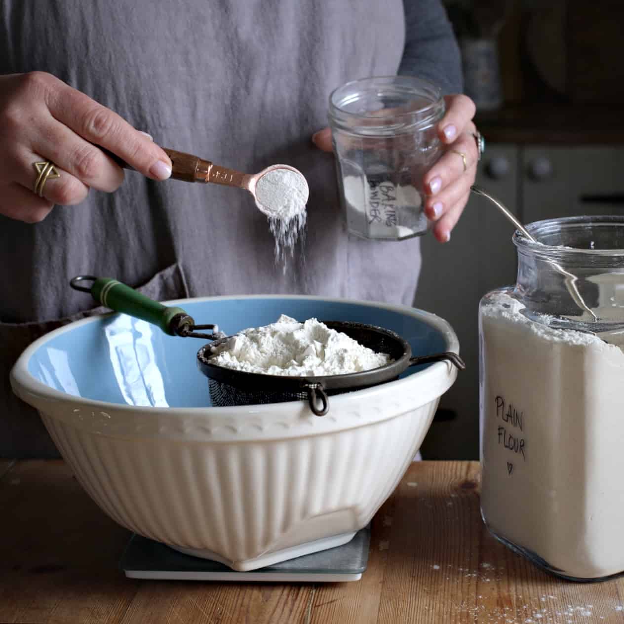 Woman’s hands tipping baking powder into an old fashioned sieve over a mixing bowl from a copper teaspoon