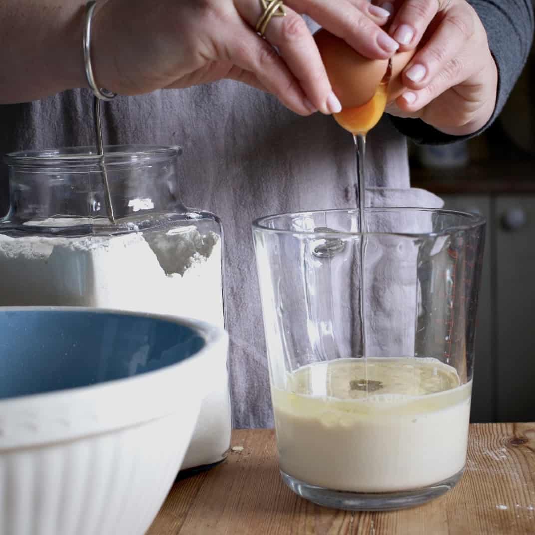 Woman cracking an egg into a glass just half filled with milk and oil on a wooden kitchen counter