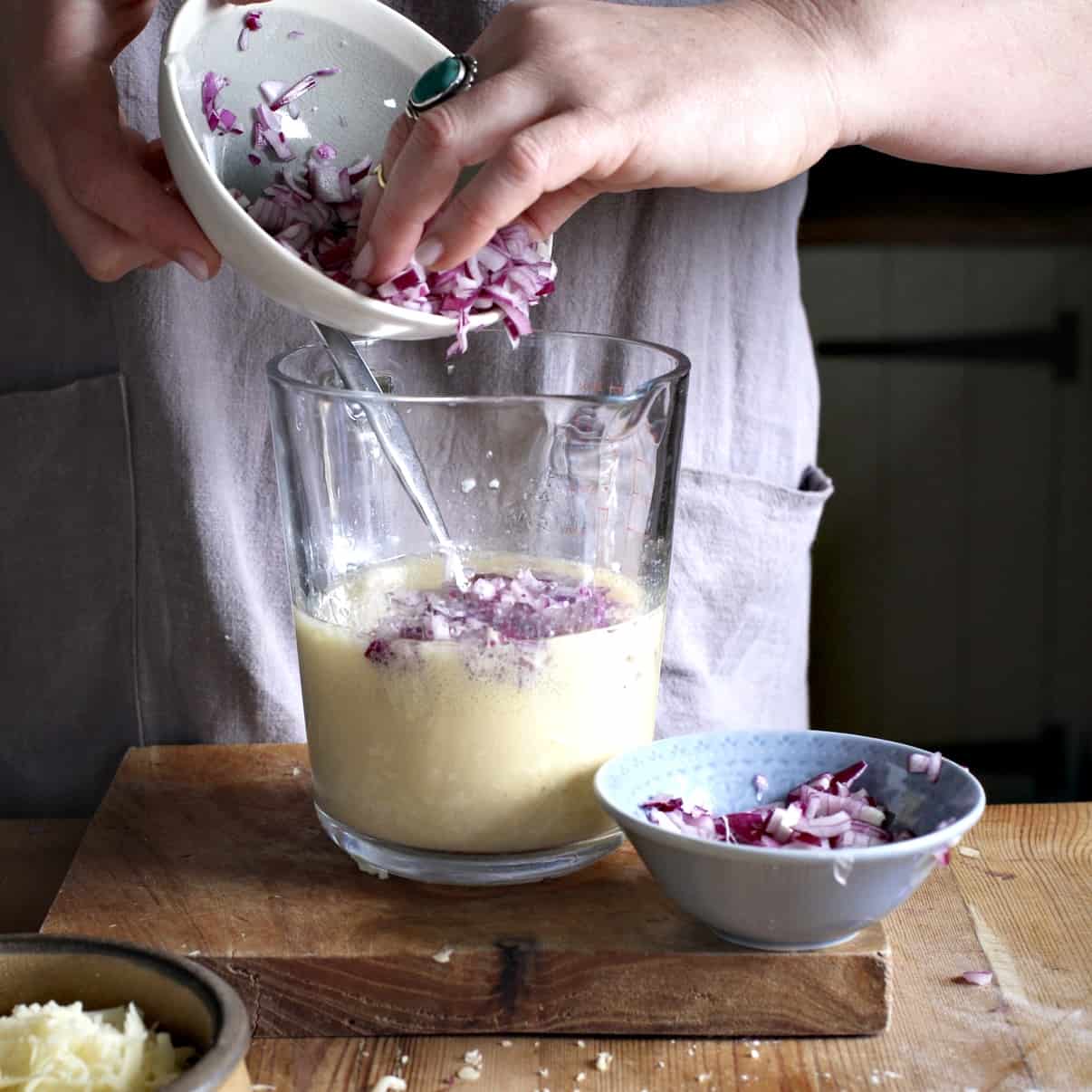 Woman in grey tipping chopped red onion from a small grey bowl into a glass jug of muffin batter