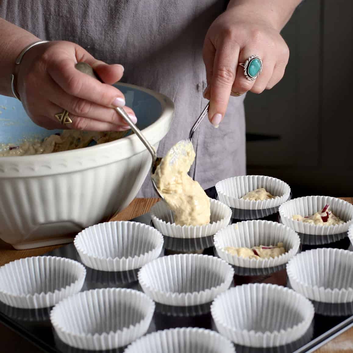 Woman’s spooning savoury muffin batter from a large white mixing bowl into white paper muffin cases