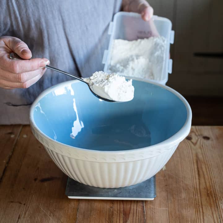 womans hands holding a metal spoon of plain flour over a large blue and white mixing bowl